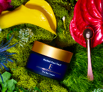 Blue and gold jar of Hyaluronic Acid skincare cream on green moss with tropical flowers and a small silver spoon.