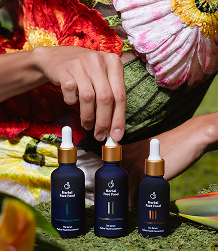 Hand adjusting three dark blue skincare dropper bottles placed on green moss with colorful flowers in the background.