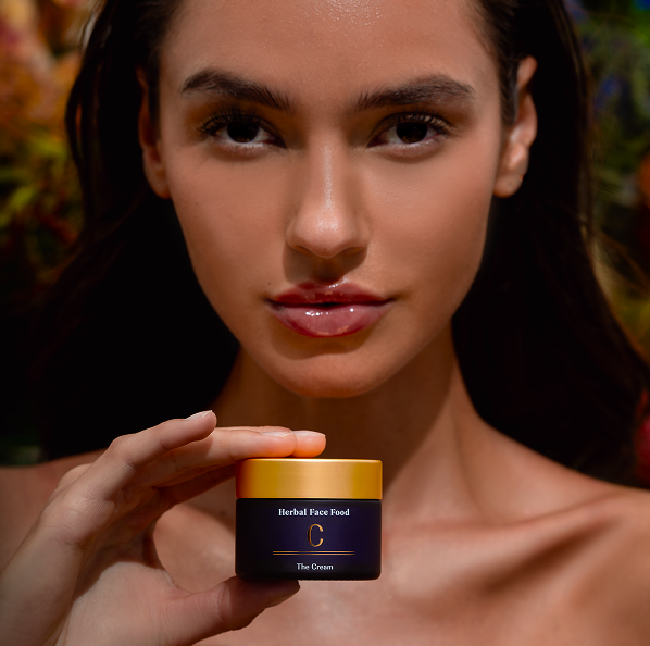 Close-up of a woman holding Herbal Face Food cream with a gold lid near her face.