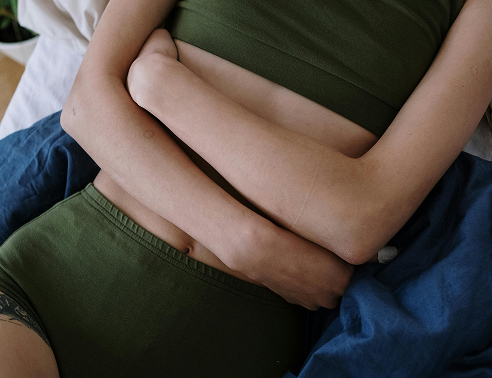 Close-up of a person wearing olive green underwear and top, lying down with arms crossed over their stomach on blue bedding.