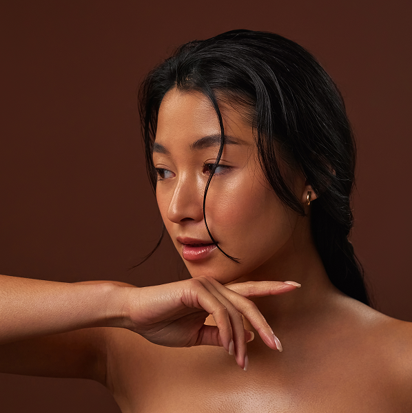 Close-up of a woman with smooth skin and dark hair holding her hand near her chin against a brown background.