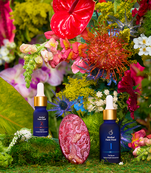 Two blue dropper bottles of Herbal Face Food serum surrounded by vibrant tropical flowers and greenery, with a decorative pink stone in the center.