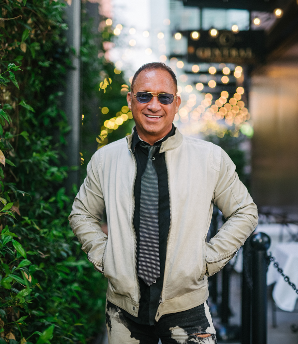 Smiling man wearing sunglasses, a light jacket, black shirt, and a patterned tie standing outdoors near green foliage with blurred lights in the background.