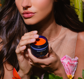 Close-up of a woman holding an open jar of vibrant orange cream with glossy pink lips and flowing brown hair.
