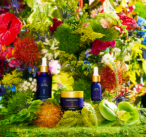 Three blue skincare product containers displayed among colorful flowers and green moss.
