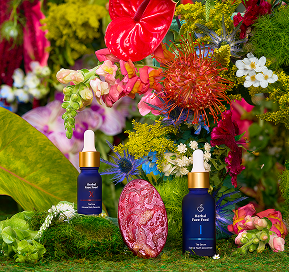 Two blue dropper bottles of skincare products displayed on green moss with diverse colorful flowers and a pink crystal.
