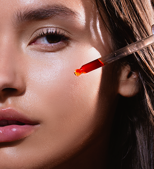 Close-up of a woman applying red serum to her cheek using a dropper.