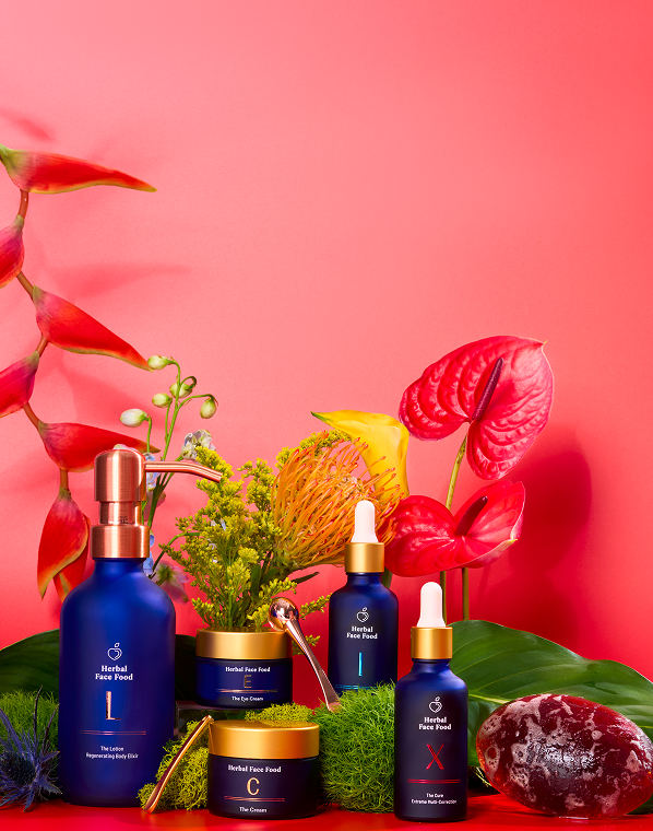 Blue bottles and jars of Herbal Face Food skincare products surrounded by colorful tropical flowers and green leaves on a vibrant pink background.
