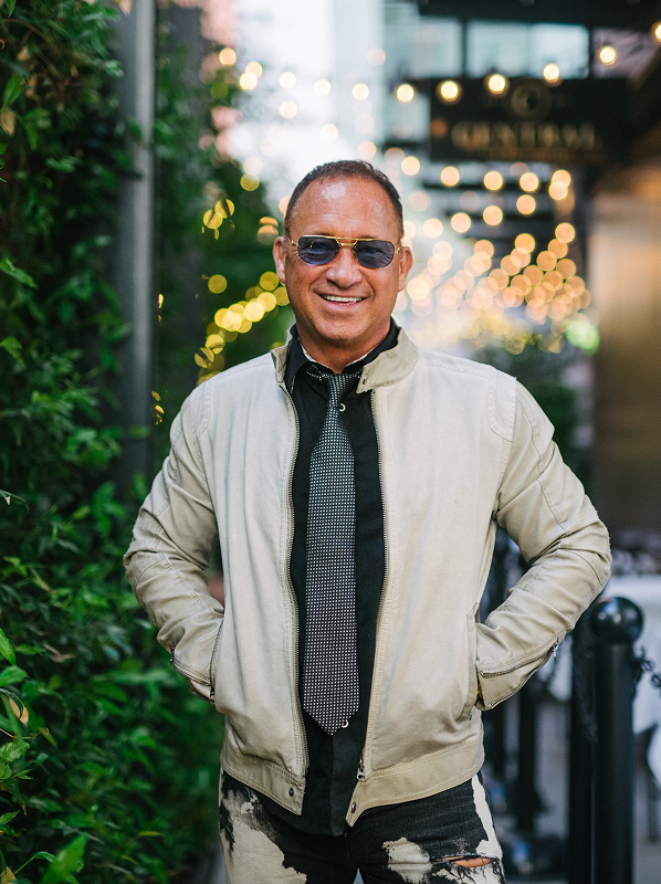 Man wearing sunglasses, a light jacket, patterned tie, and distressed jeans standing outdoors with hands in pockets and bokeh lights in background.