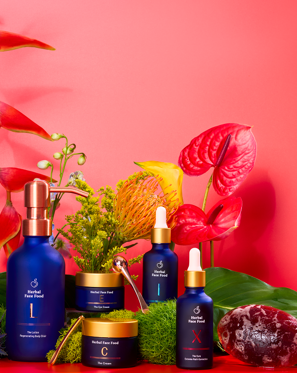Blue and gold Herbal Face Food skincare bottles and jars arranged with vibrant tropical flowers and greenery against a pink background.
