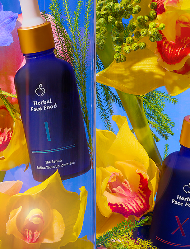 Two blue bottles of Herbal Face Food serum surrounded by yellow and pink orchids and greenery against a bright blue background.
