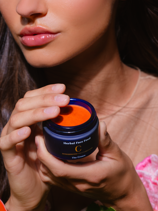 Close-up of a woman holding and touching an open blue jar of orange herbal face cream labeled 'Herbal Face Food C The Cream.'