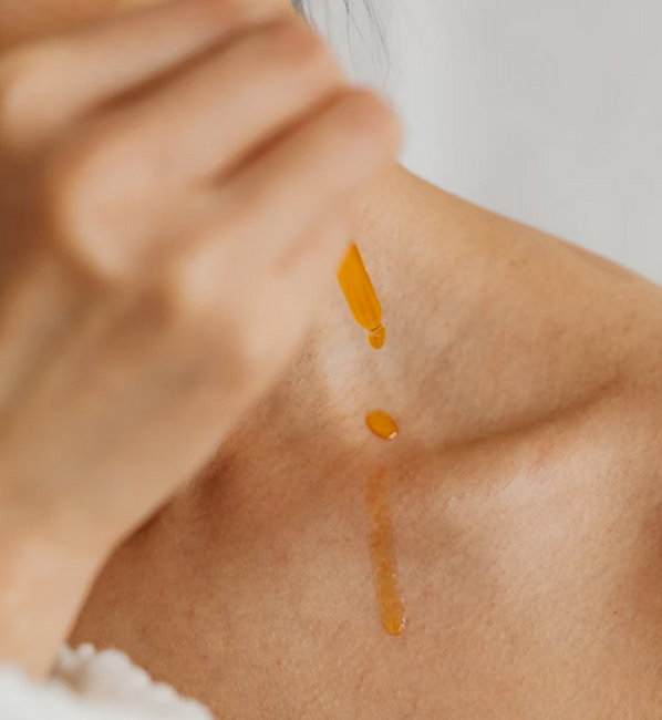 Dropper releasing golden liquid drops onto a person's collarbone skin.