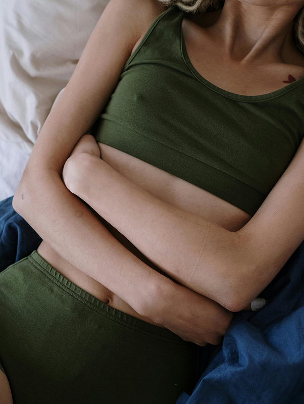 Person lying down on a bed with arms crossed over abdomen wearing olive green sleeveless top and matching bottoms.
