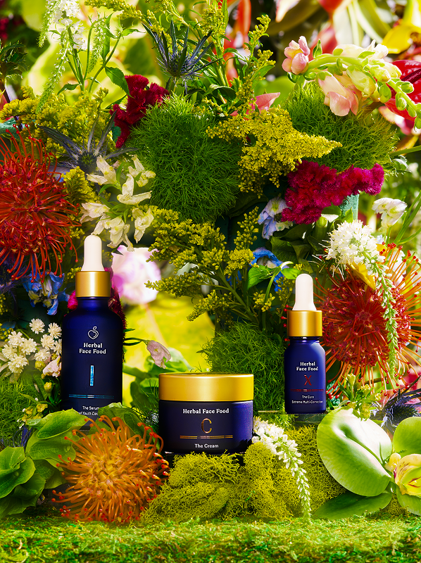 Three Herbal Face Food skincare products with blue and gold packaging displayed among vibrant, colorful flowers and green foliage.