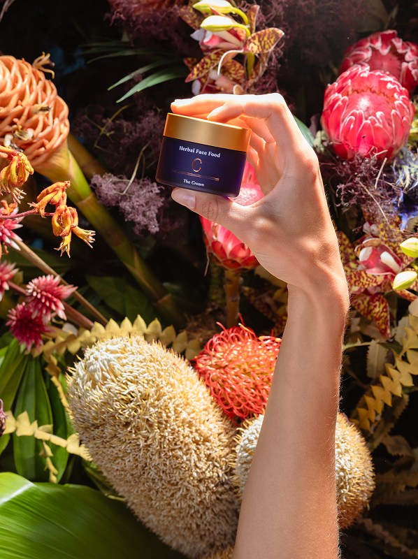 Hand holding a jar of Herbal Face Food cream against a background of colorful exotic flowers and foliage.
