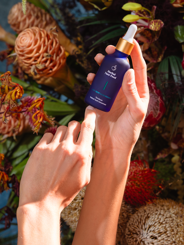 Two hands holding a blue dropper bottle labeled Herbal Face Food against a background of various flowers and plants.