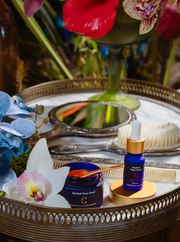 A vintage tray holding herbal face food skincare jars with a gold spoon, a hand mirror, a white comb, a brush, and orchid flowers.