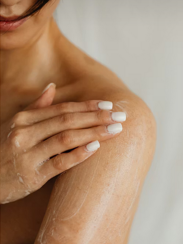 Close-up of a person applying lotion on their shoulder with manicured white nails.
