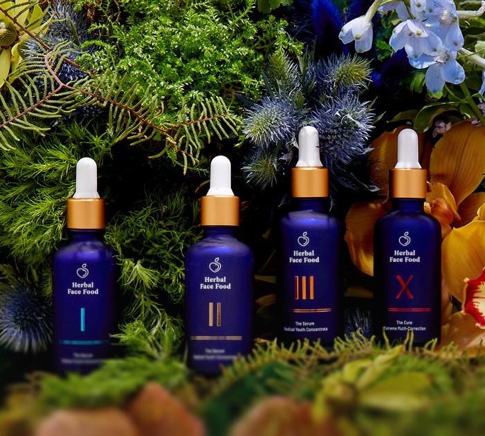 Four blue dropper bottles of Herbal Face Food skincare products arranged among green foliage and flowers.