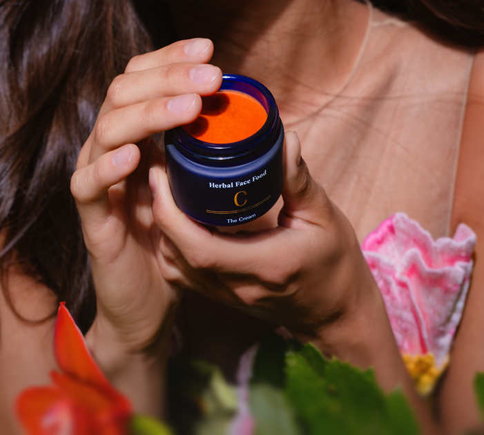 Person holding an open blue jar of bright orange herbal face cream labeled 'Herbal Face Food C The Cream'.