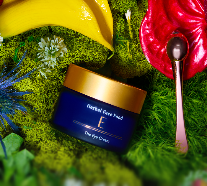 Blue jar of Herbal Face Food E the Eye Cream with a gold lid, surrounded by green moss, yellow banana-shaped leaves, small white flowers, a red decorative leaf, and a spoon with a dark rounded tip.