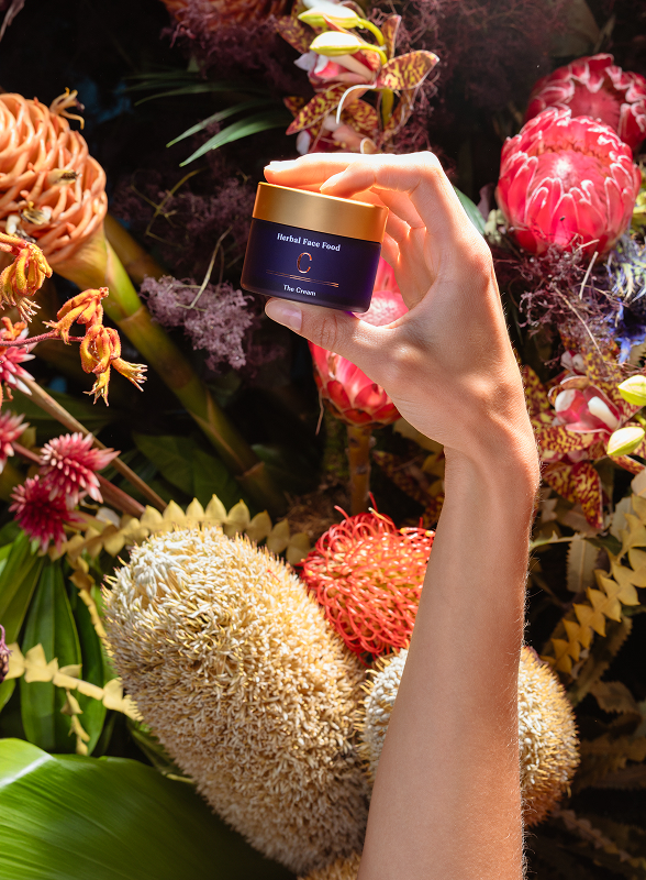 Hand holding a small jar of Herbal Face Food cream above vibrant tropical flowers and green leaves.