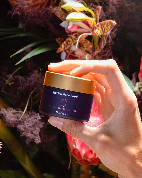 Hand holding a jar of Herbal Face Food cream with a golden lid against a background of flowers and foliage.