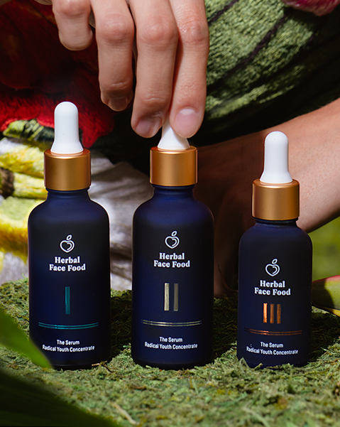Three dark blue bottles of Herbal Face Food serum with gold caps and white droppers on green moss, one held by a hand.