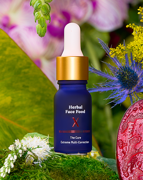 Blue glass dropper bottle with gold cap labeled 'Herbal Face Food The Cure Extreme Multi-Correction' surrounded by green leaves and colorful flowers.