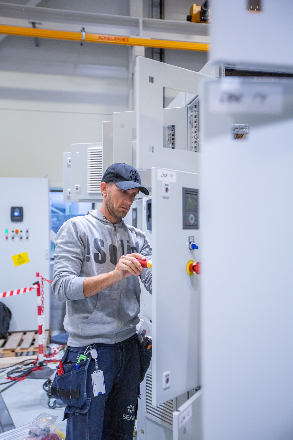 Technician wearing a cap and gray hoodie using a screwdriver to work on an industrial control panel in a factory setting.