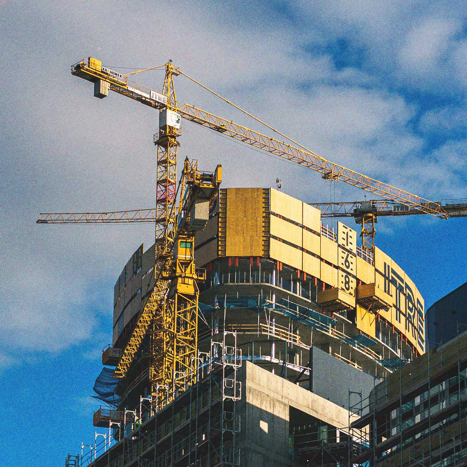 High-rise building under construction with scaffolding, concrete structure, and two yellow tower cranes against a partly cloudy blue sky.