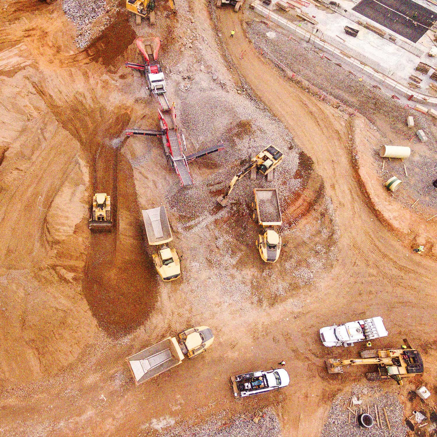 Aerial view of a construction site with multiple yellow dump trucks, excavators, a conveyor system, and two white pickup trucks on dirt and gravel terrain.