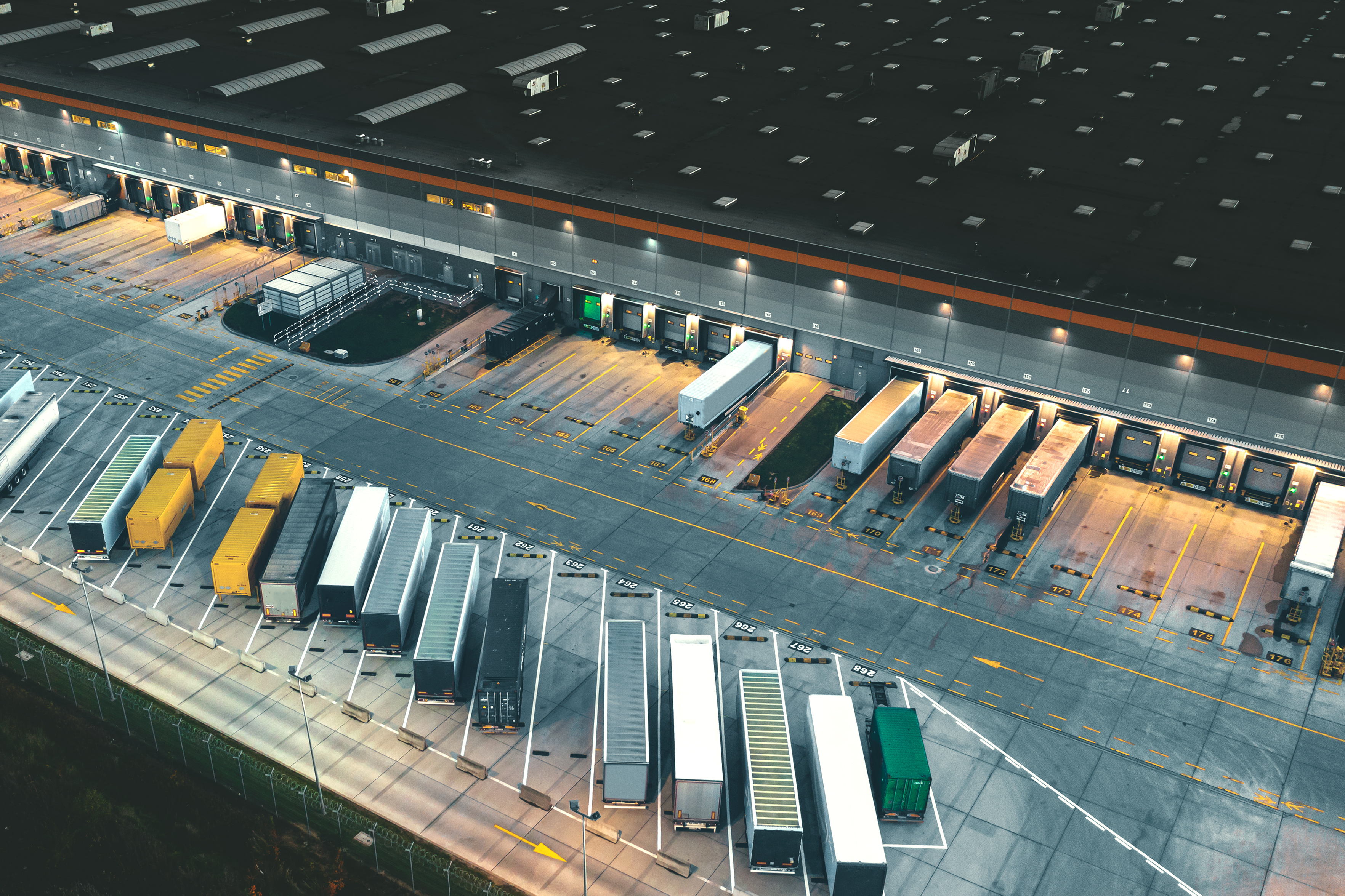 Aerial view of a modern warehouse with numerous semi-trailers and trucks parked at loading docks and in a designated parking area.