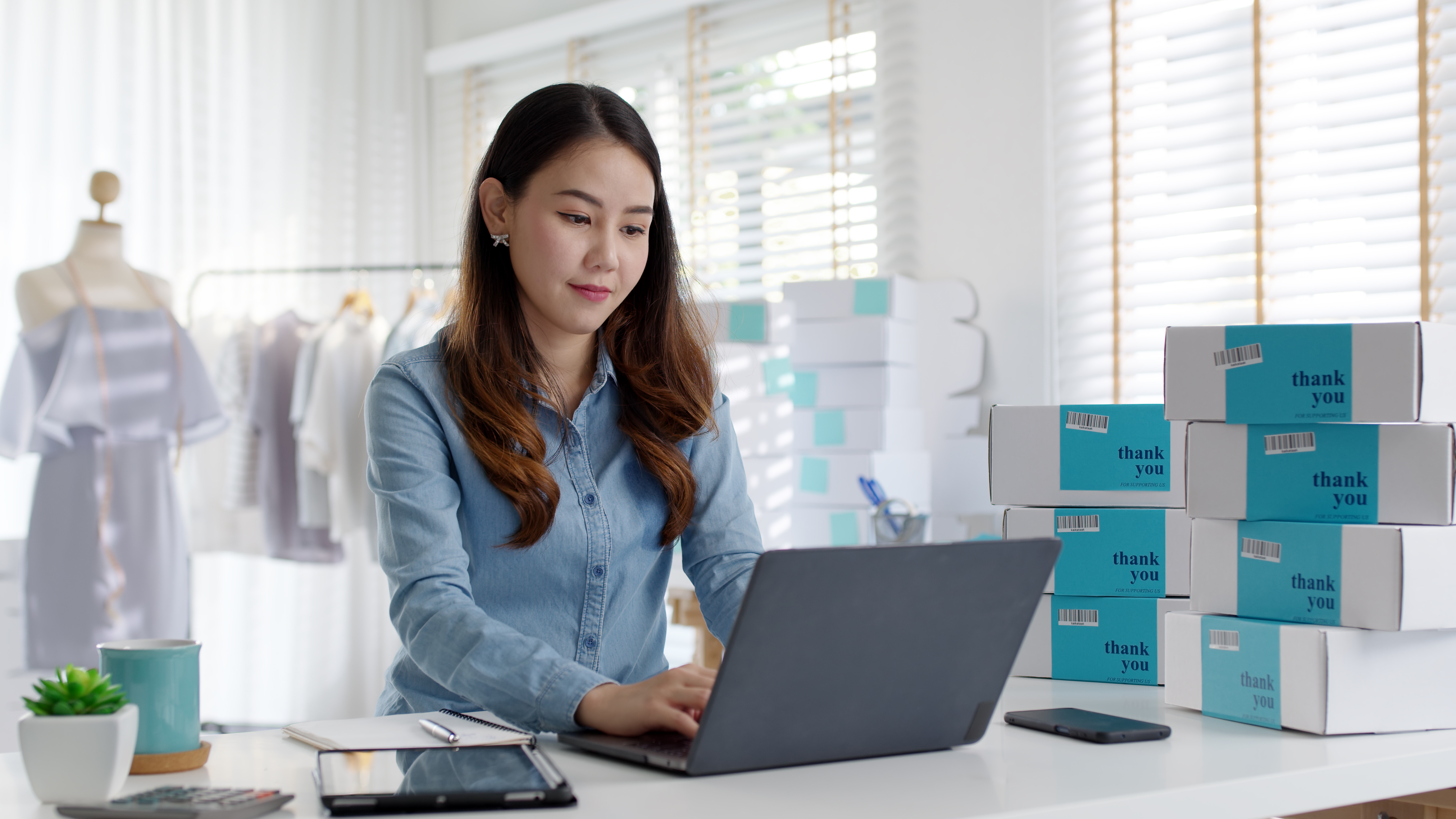 Young woman working on a laptop at a desk surrounded by stacked thank you labeled shipping boxes in a bright room.