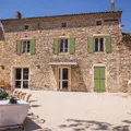 Stone facade of a house with green shutters and a sunny terrace with white chairs.