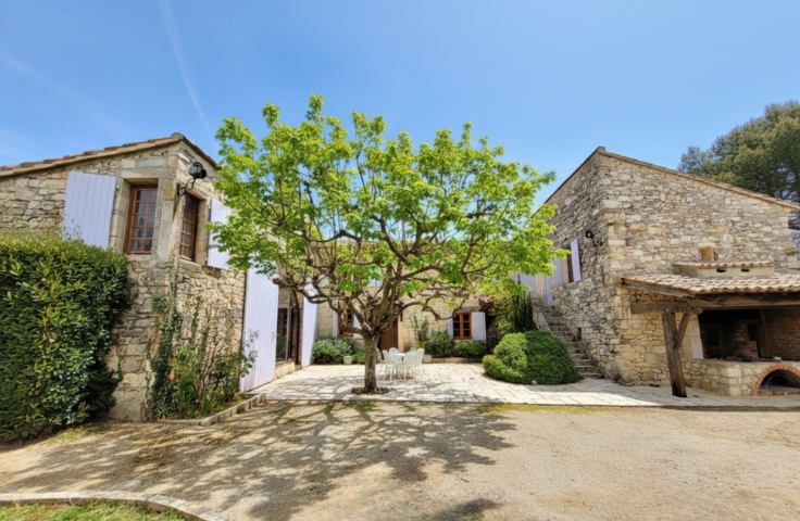 Extérieur d'un mas provençal en pierre, cour ensoleillée, grand arbre feuillu au centre, table et chaises blanches sous l'arbre, volets clairs lavande, escalier extérieur en pierre, four à pizza ou à pain sous un auvent
