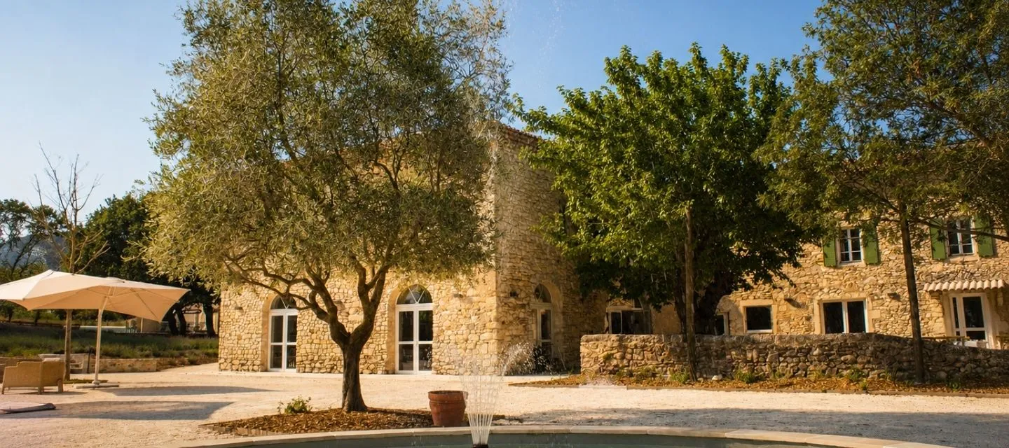 Entrée du Domaine de la Vivande, mas provençal en pierre et toits en tuiles, fontaine octogonale avec jet d'eau au premier plan, bassin entouré de lavande en fleurs