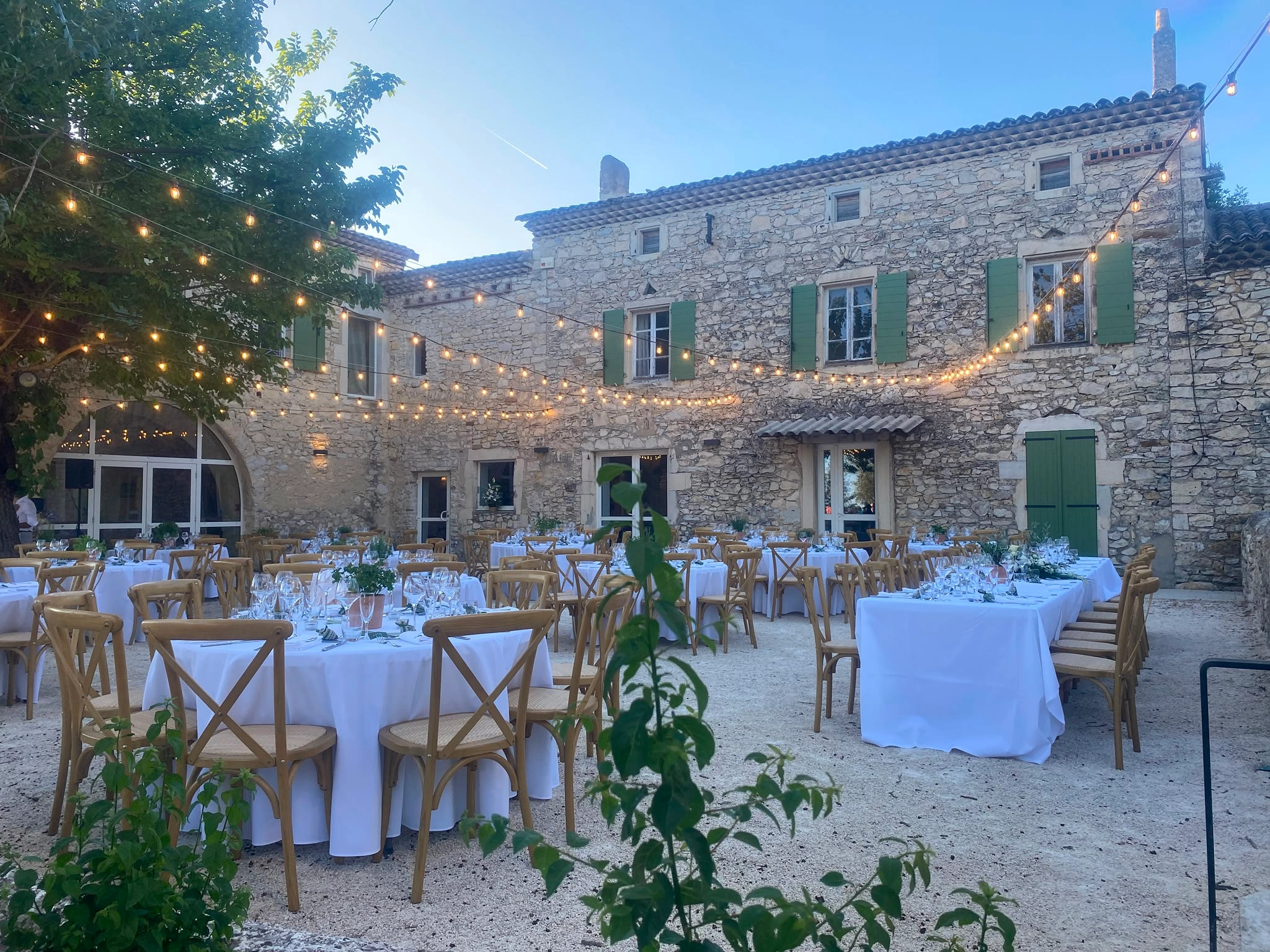 Terrasse provençale et façade de mas en pierre, salons de jardin en rotin avec coussins gris, grands pots en terre cuite remplis de lavande, olivier à gauche, pelouse et paysage de campagne
