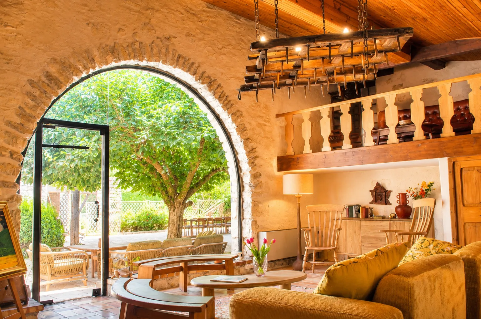 Rustic Provençal farmhouse style living room, large stone arch opening onto the outside, beige corner sofa, antique green armchair, stone fireplace, wooden mezzanine with bookcase