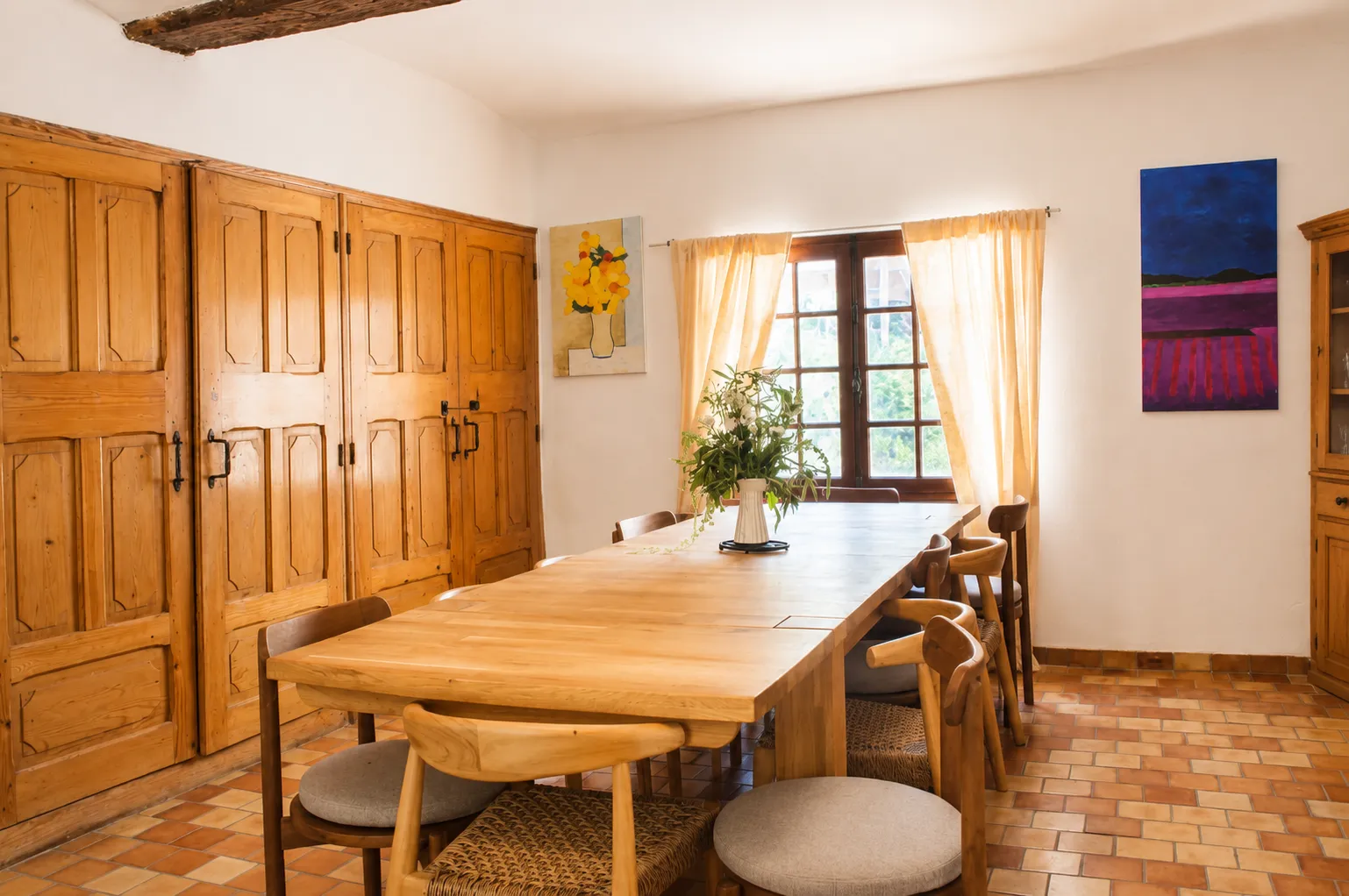Contemporary Mediterranean style dining room and living room, vaulted ceilings, light walls, light wood dining table, white bookcase, archway opening onto the living room.