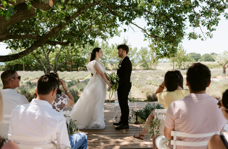 Organisation d'un mariage au Domaine de la Vivande, mariés marchant dans l'allée sous une pluie de confettis ou de pétales, invités applaudissant, cérémonie extérieure, allée bordée de pots de lavande et de chaises croisées en bois