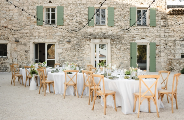 Espace dîner plein air, tables rondes dressées avec nappes blanches, guirlandes lumineuses suspendues au-dessus d'un grand arbre et de la terrasse de la bastide
