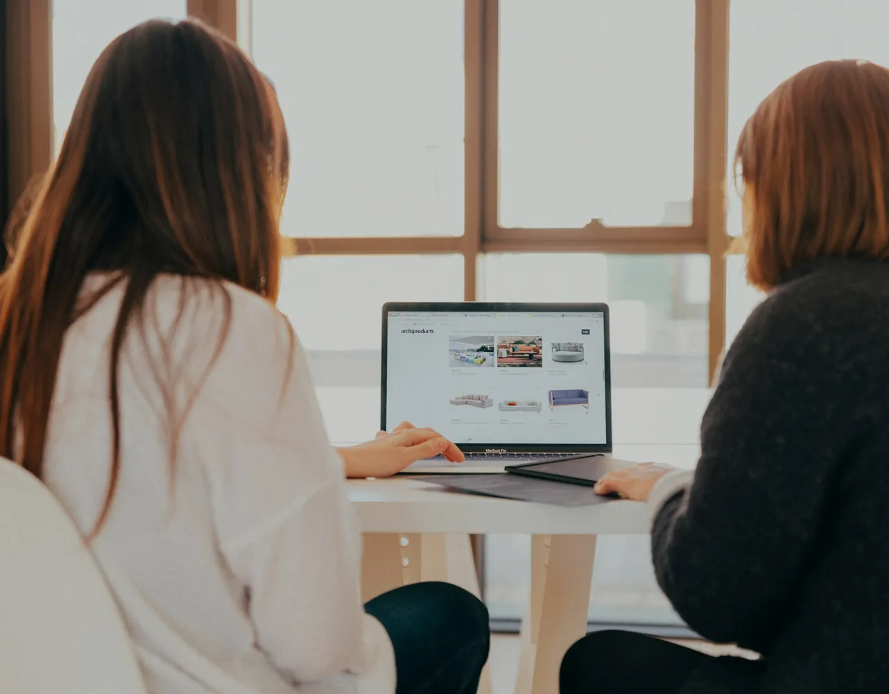 Two women seated at a table looking at a laptop screen displaying images of furniture products.