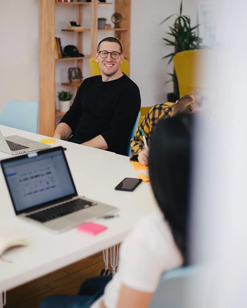 Man wearing glasses and a black shirt smiling at a meeting table with laptops, a mobile phone, and people working.
