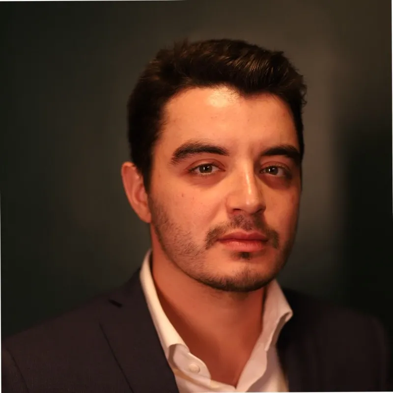Portrait of a man with dark hair and light facial hair wearing a dark suit jacket and white shirt against a dark background.