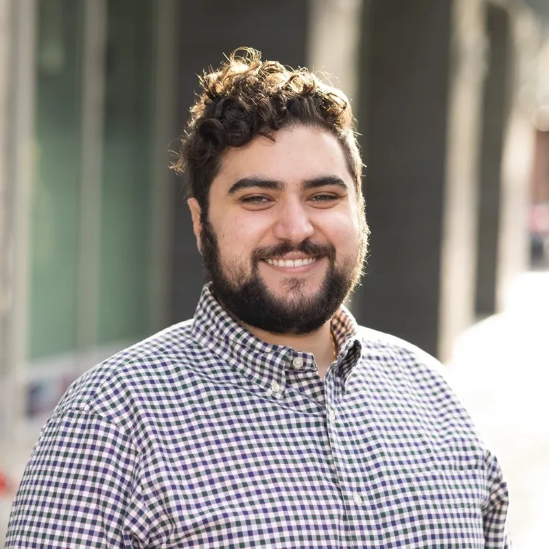 Smiling man with curly hair and beard wearing a checkered button-up shirt outdoors.
