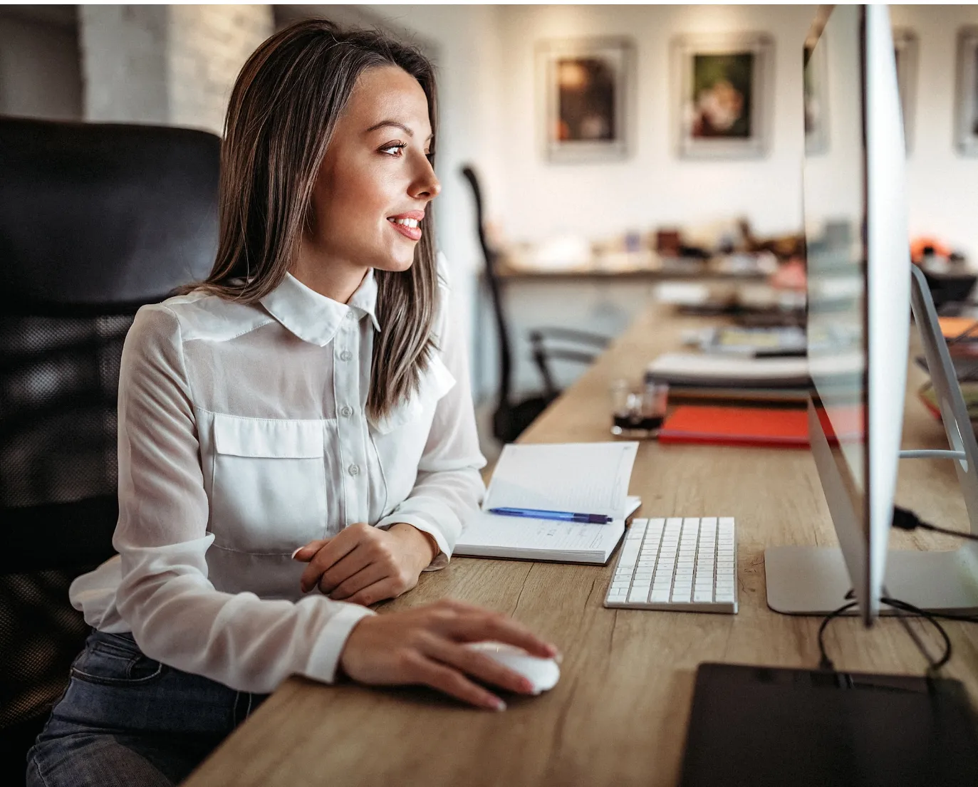 Smiling woman in white blouse using a computer mouse at a desk with a keyboard and notebook.