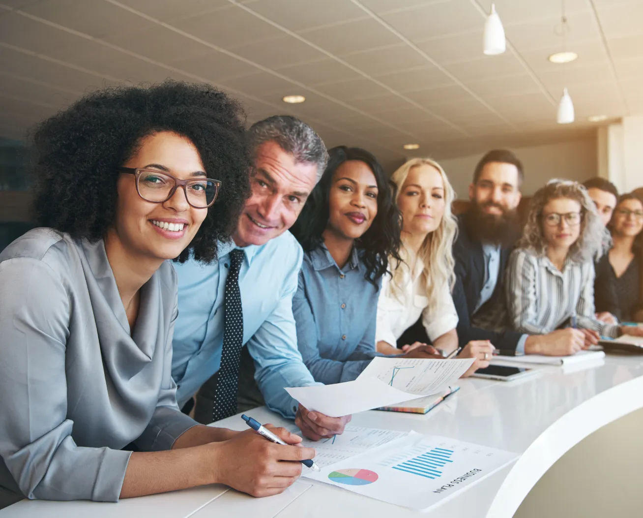 A diverse group of seven professionals sitting at a curved table, reviewing business documents and charts.