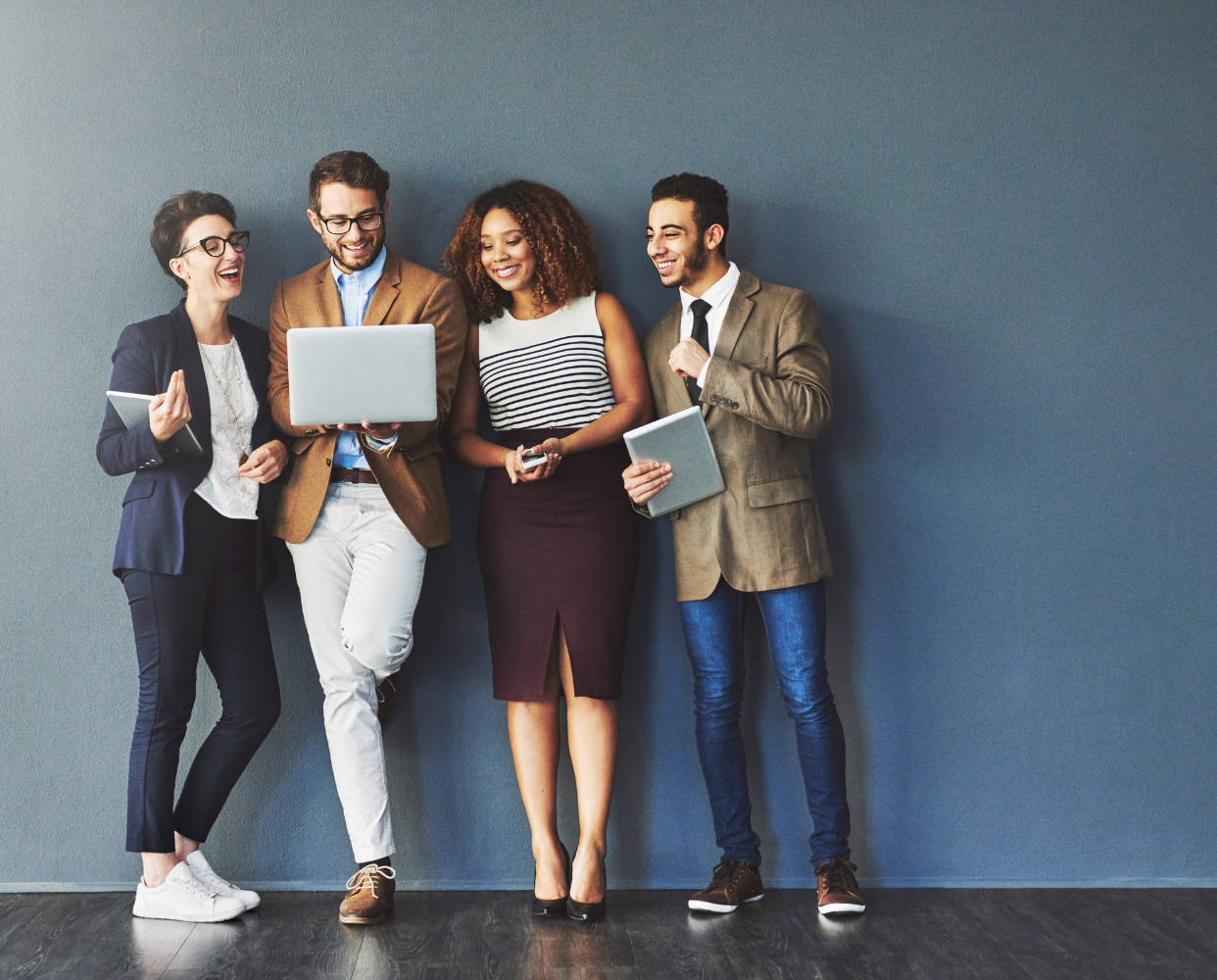 Four diverse business professionals smiling and collaborating while looking at a laptop and holding digital devices against a gray wall.